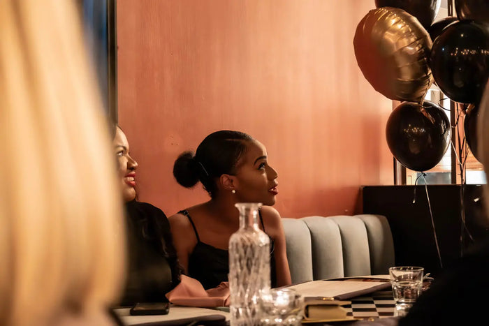 Two women sitting in a restaurant booth look up at balloons, smiling. 
