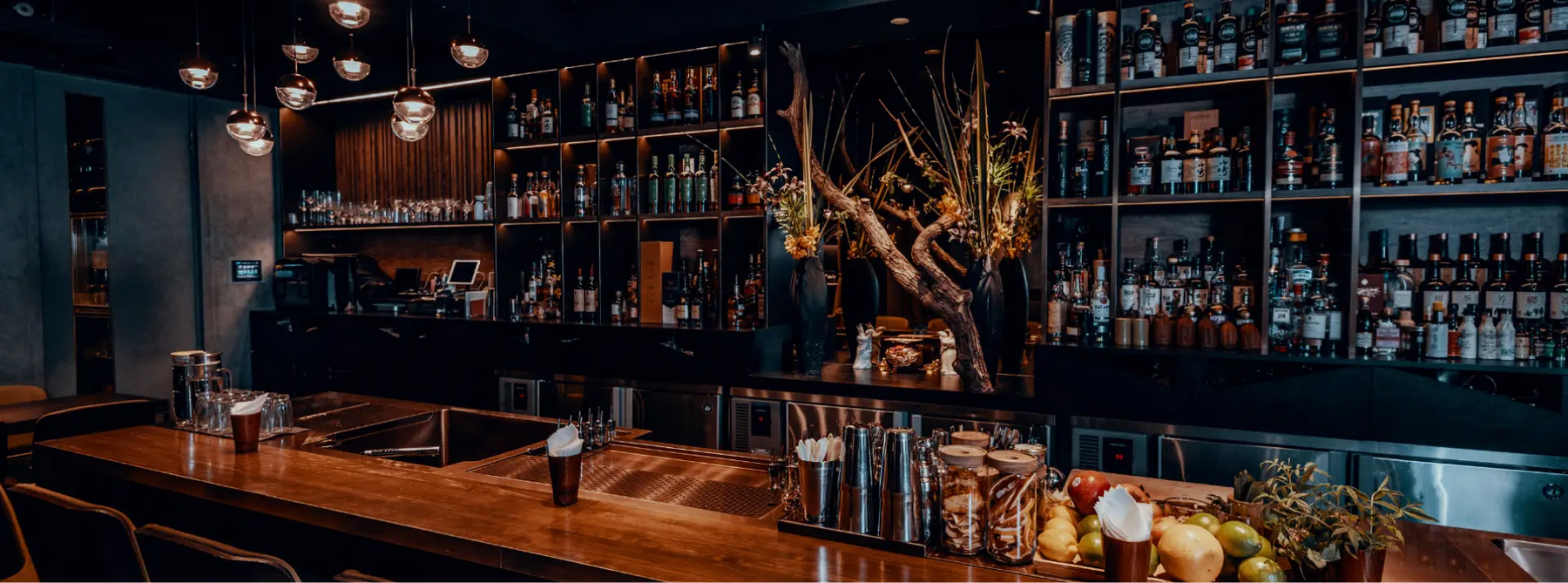 A dimly lit, fully equipped bar with a wooden counter, leathered stools and an assortment of bar equipment