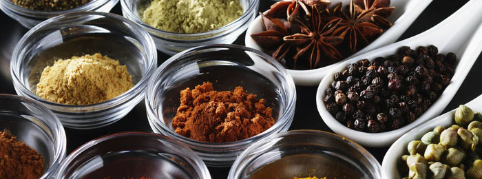 Collection of whole and ground spices in glass bowls and ceramic spoons on a dark background.