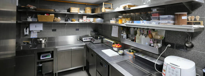 Small commercial kitchen with stainless steel counters, wall-mounted shelves, compact appliances, and organized prep stations.