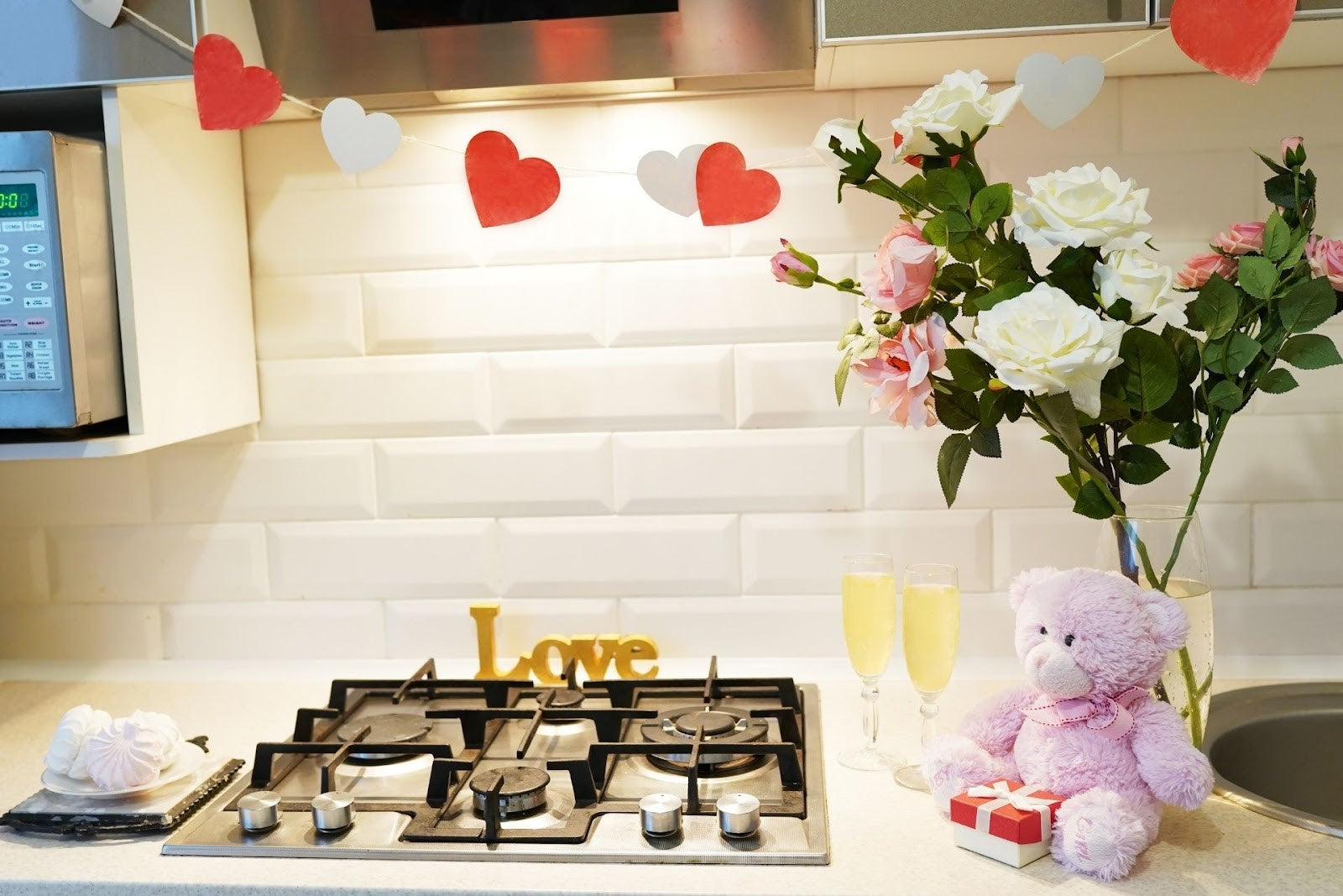 Paper hearts, flowers, a teddy bear and two glasses of champagne on a kitchen counter next to a stovetop