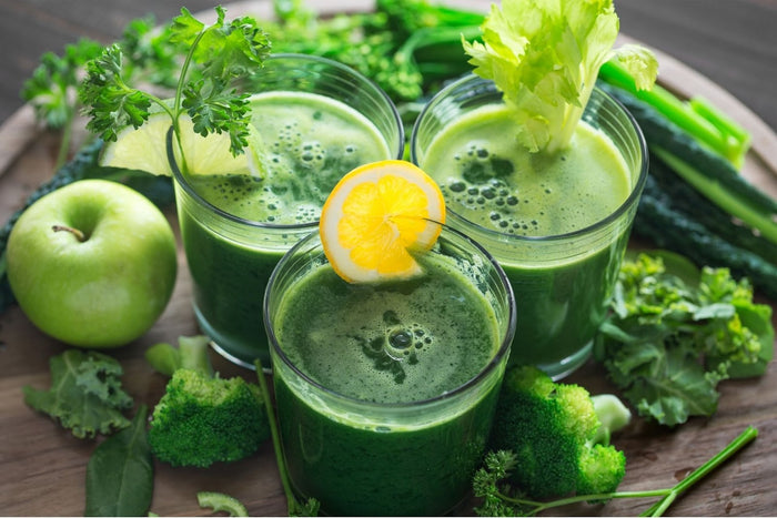 Three glasses of green juice cleanses surrounded by a green fruits and vegetables