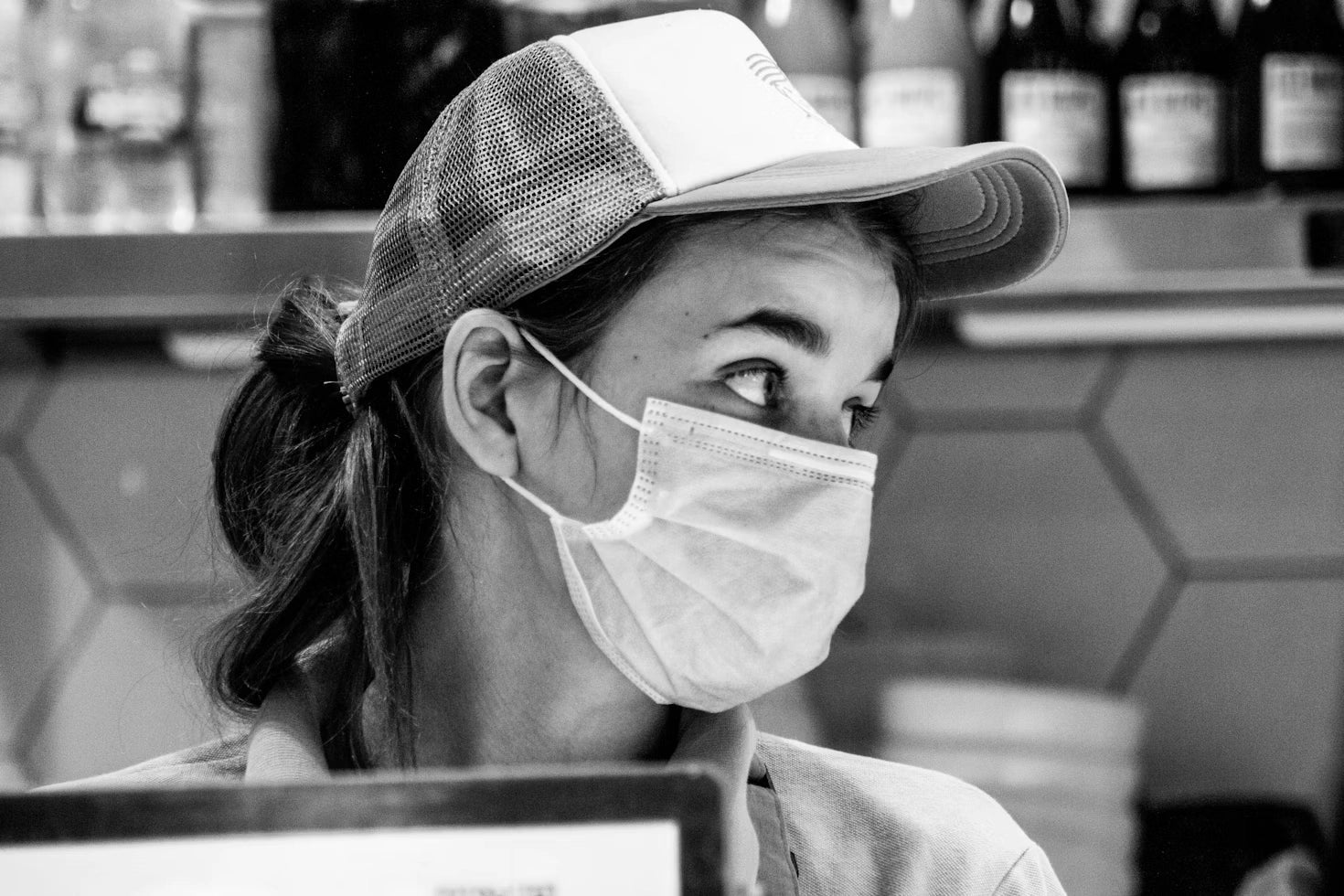 A greyscale image of a female food cashier wearing a cap and mask, looking sideways. 