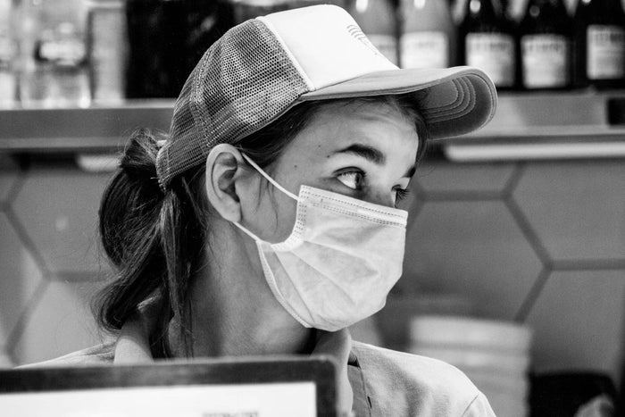 A greyscale image of a female food cashier wearing a cap and mask, looking sideways. 