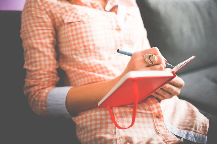 A woman wearing orange plaid writes in her red notebook. 