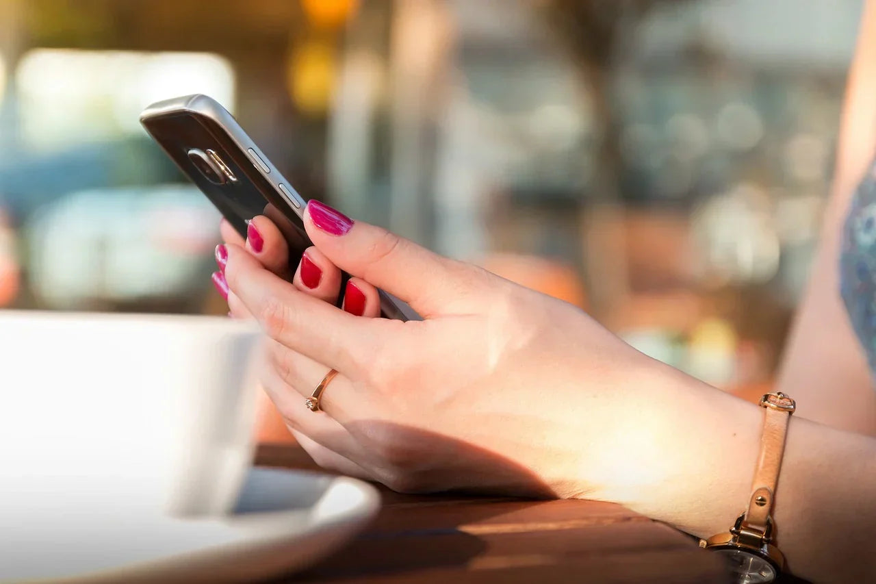 A woman's hand holds a smartphone next a to a white coffee cup. 