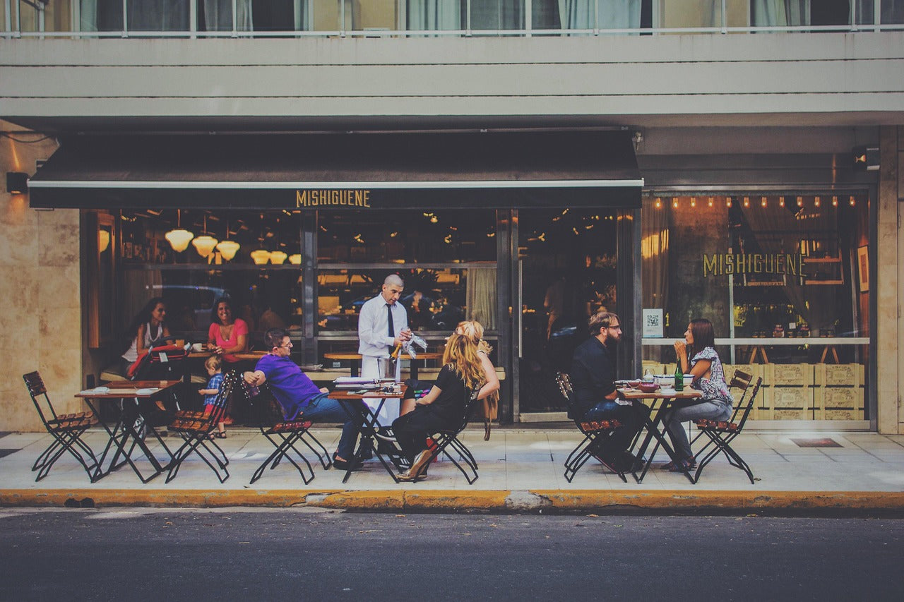 Curbside view of a restaurant outdoor patio with seated guests and a waiter. 