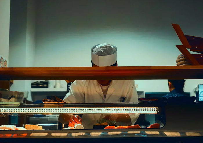 A chef, whose face is covered by a wooden slab, garnishes sushi under countertop lighting. 