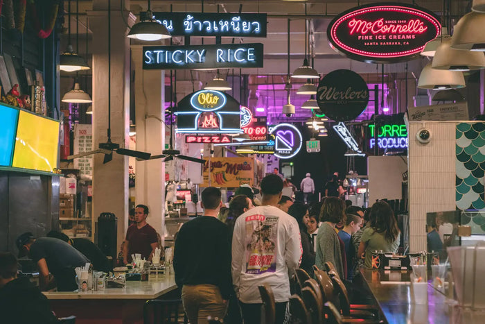 Food market is filled with customers and restaurant signage hanging above them. 