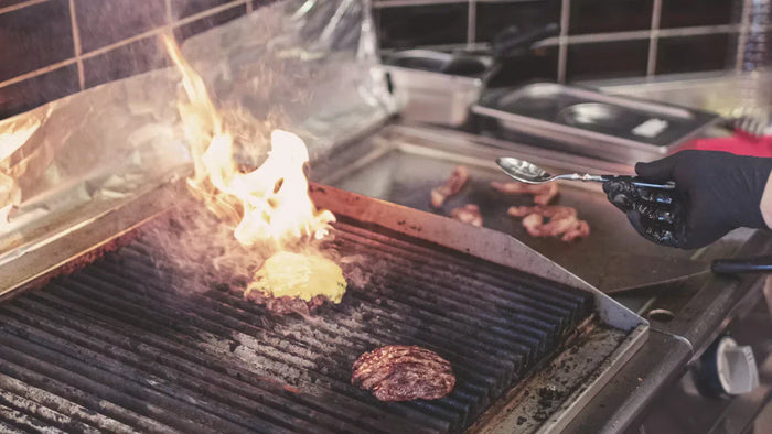 Close up of a BBQ grill with burger patties, one catching a flame while a cook's hand hovers nearby.