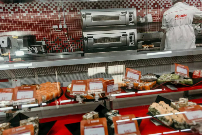 An enclosed glass counter in front of kitchen ovens displays sushi and spring rolls. 