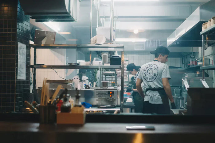 Three line cooks work next to grills, refrigerators and vents in a well-lit kitchen space.