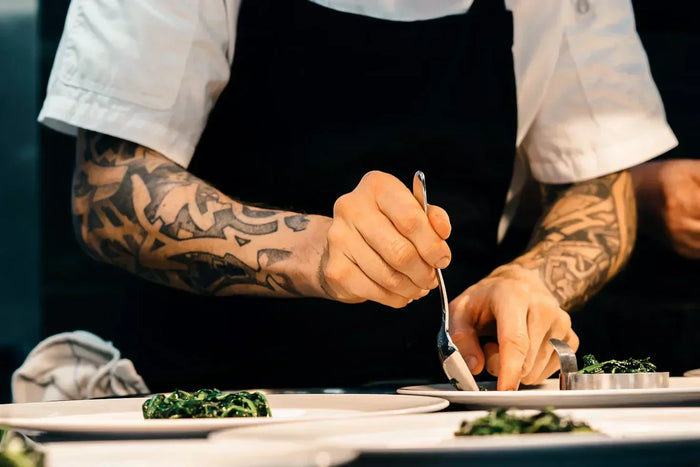 A tattooed cook wearing a black apron plates a veggie dish onto a plate. 