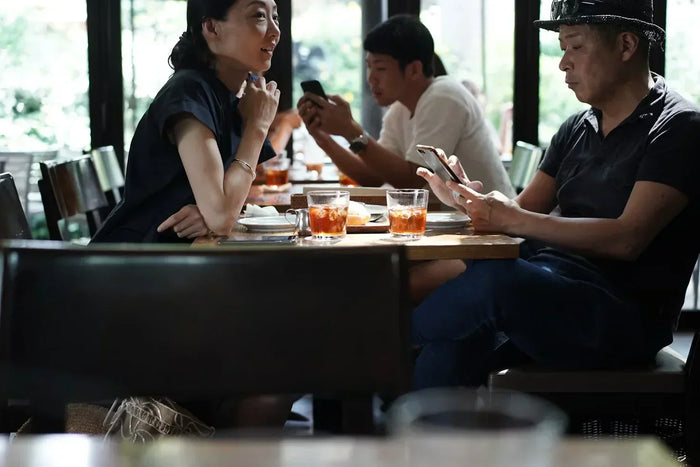 A wife seated at a table looks afar, while the husband is on his phone. 