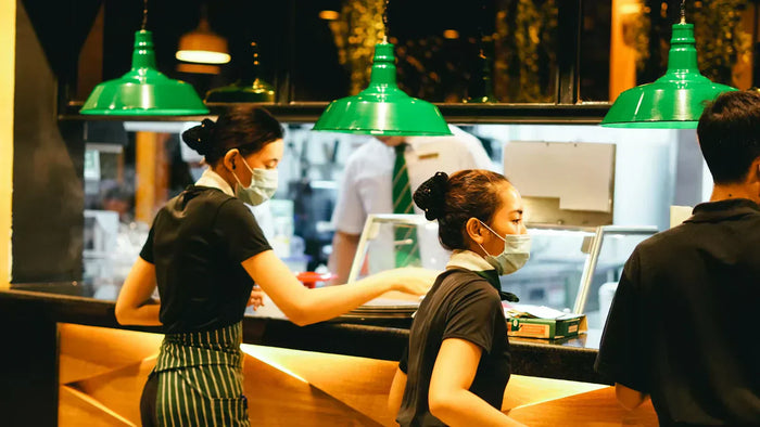 Two female restaurant employees wearing masks and black tees work beside a kitchen counter.