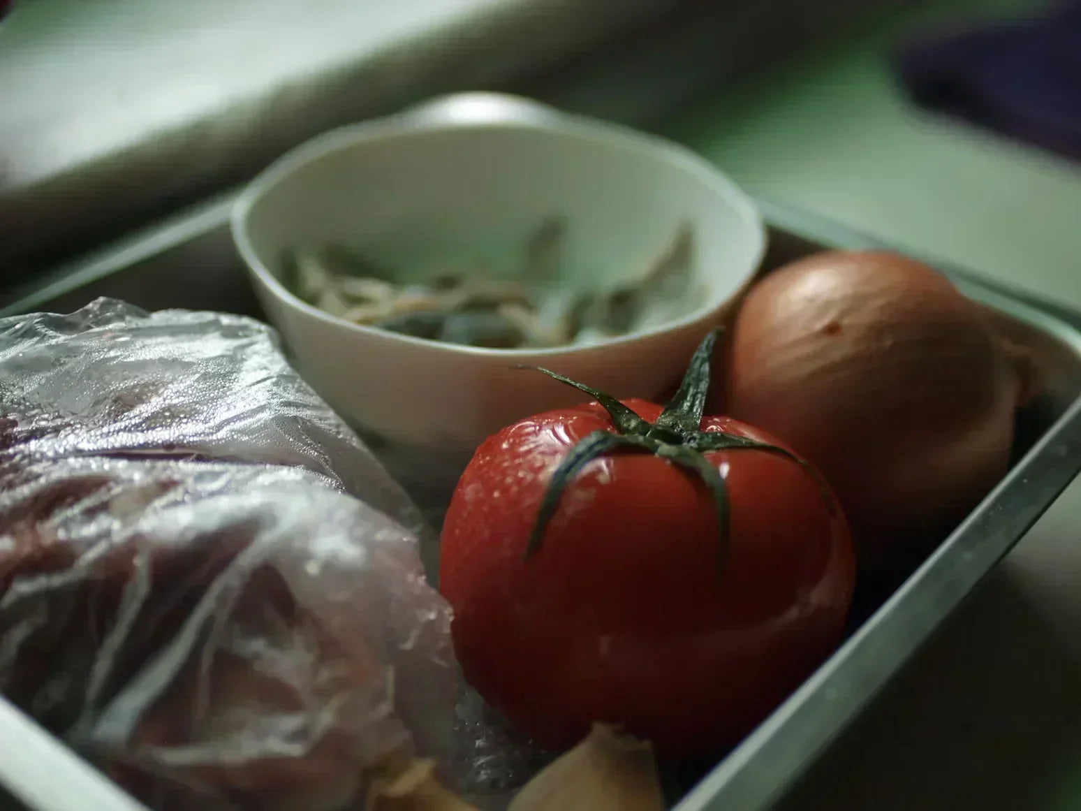 A tray contains a slightly mushy tomato, an onion, plastic-wrapped food, and a veggie bowl. 