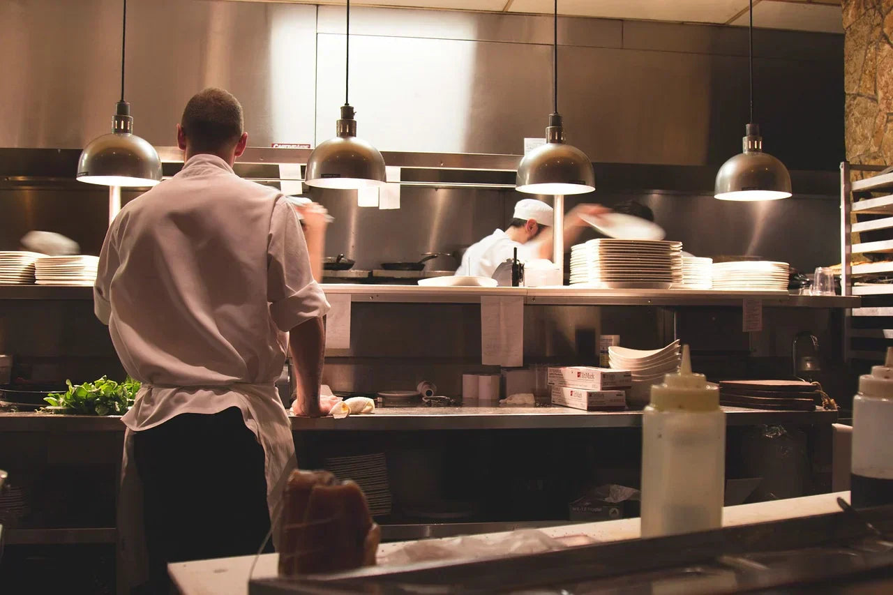 Line cooks prepare foods on silver counter tops under soft-lit hanging kitchen fixtures. 
