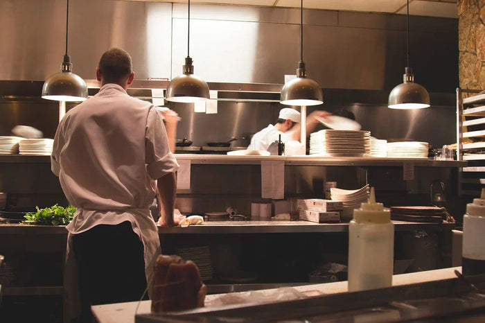 Line cooks prepare foods on silver counter tops under soft-lit hanging kitchen fixtures. 